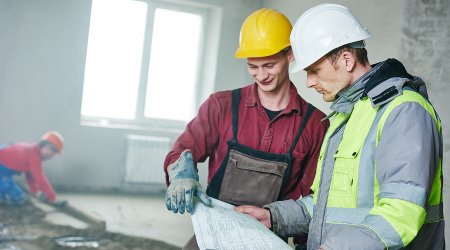 Two men wearing hard hats looking at building plans. one is wearing a red shirt with a grey apron and gloves and the other is wearing a grey and yellow high visibility jacket.