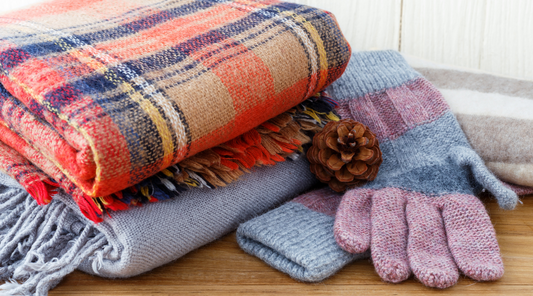 A cosy display of folded woollen blankets and knitted gloves, with a pinecone for decoration.